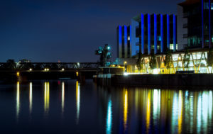 Nachtaufnahme eines Gebäudes mit blauen Lichtern und reflektierenden Wasseroberfläche in einem urbanen Umfeld.