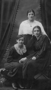 Drei Frauen in historischen Kleidern, posierend vor einem dunklen Hintergrund mit dekorativem Vorhang.